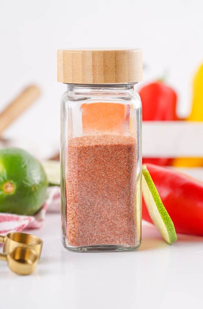 A glass jar with a wooden lid filled with a reddish-brown spice blend, likely homemade Tajin seasoning, is placed on a white surface with assorted vegetables and a brass spoon in the background.