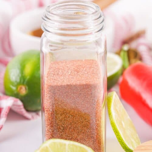 Clear spice jar filled with reddish Tajin-inspired seasoning, surrounded by sliced limes, a whole lime, red chili pepper, and kitchen utensils on a white surface with a striped cloth in the background.