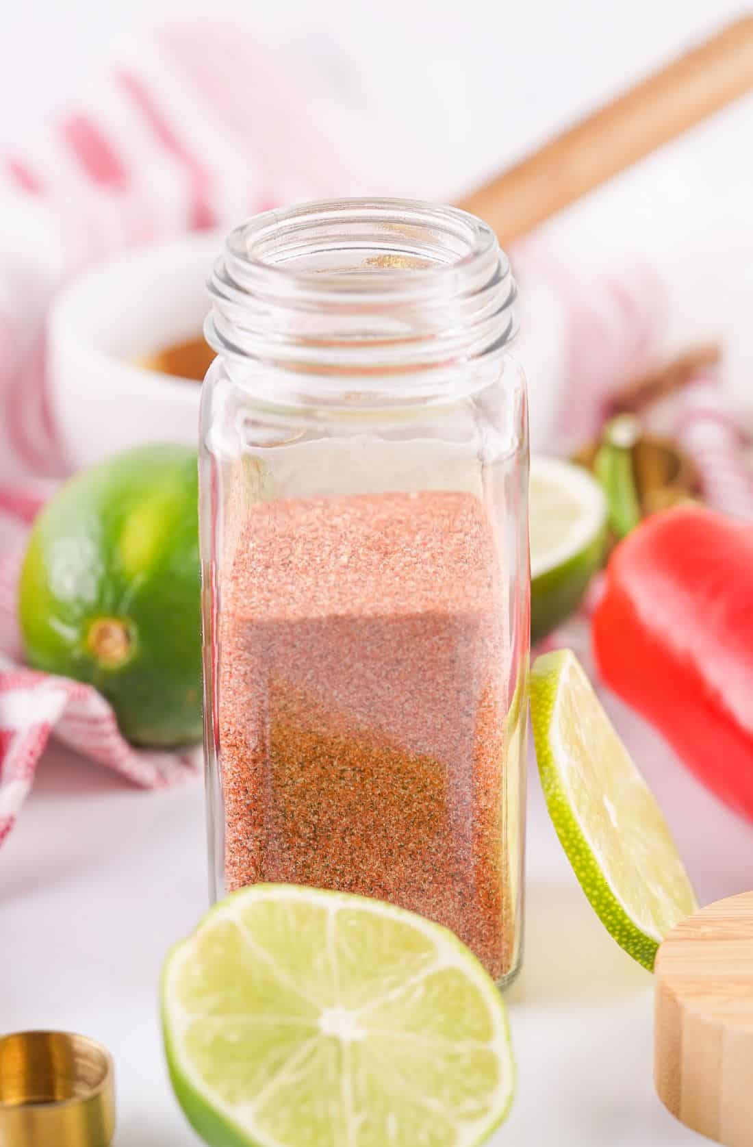Clear spice jar filled with reddish Tajin-inspired seasoning, surrounded by sliced limes, a whole lime, red chili pepper, and kitchen utensils on a white surface with a striped cloth in the background.