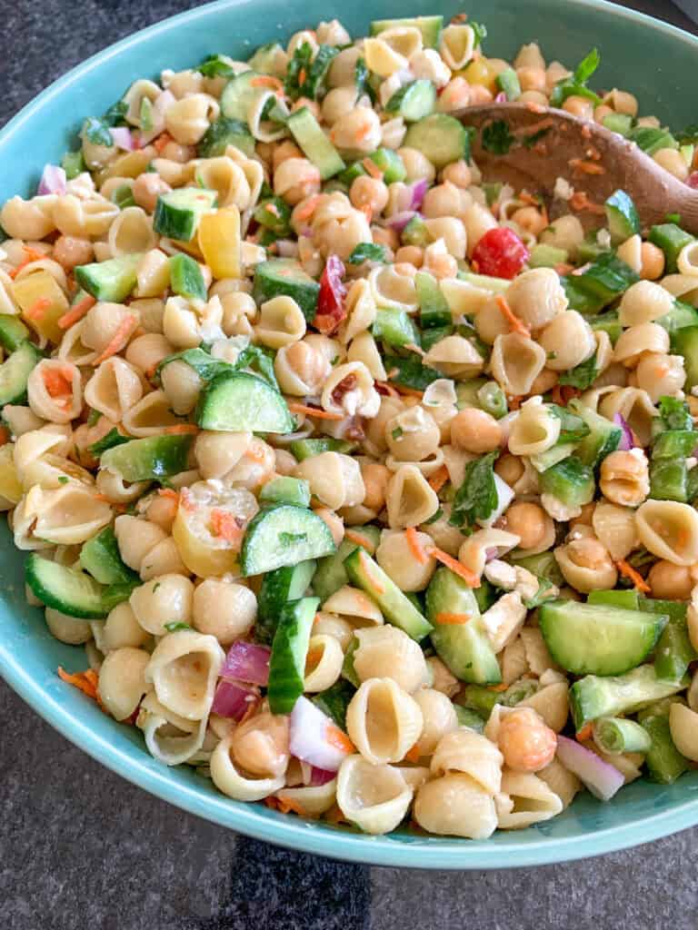 Lemon Tahini Pasta Salad 7 A bowl of Lemon Tahini Pasta Salad with shell pasta, mixed vegetables including cucumbers, red onions, peppers, chickpeas, and fresh herbs, stirred with a wooden spoon.