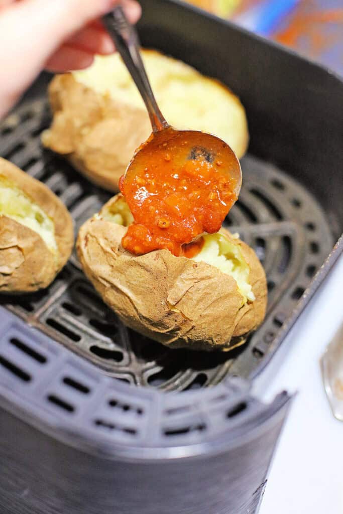 A hand is spooning chili cheese sauce onto a baked potato inside an air fryer, with other baked potatoes in the background.