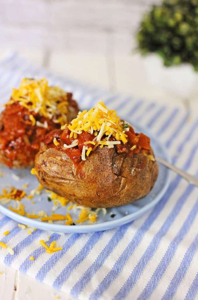 A chili cheese baked potato topped with grated cheese and tomato sauce is served on a light blue plate with a striped cloth underneath.