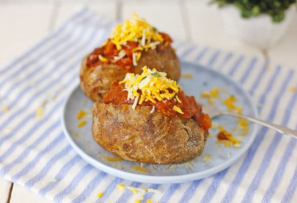 Two Air Fryer Chili Cheese Baked Potatoes on a blue plate topped with savory chili and melted cheese, all set against a charming striped cloth background.