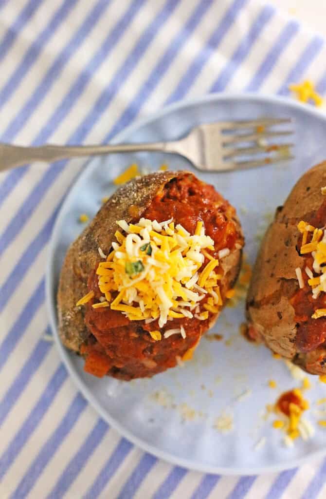 Two chili cheese baked potatoes topped with shredded cheese sit on a blue plate, which is placed on a blue and white striped cloth. A fork is positioned next to the plate.