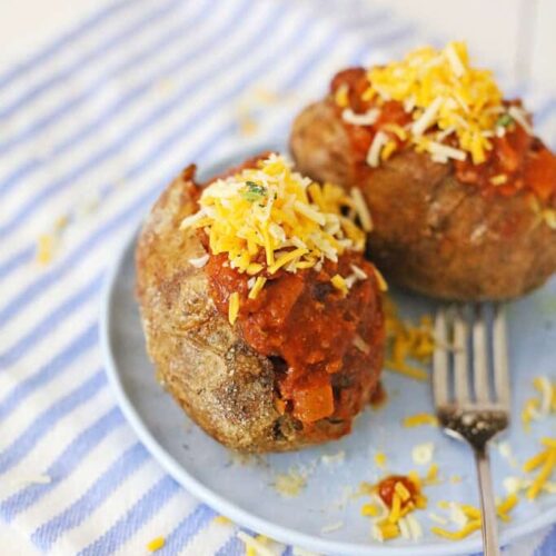 A duo of air fryer chili cheese baked potatoes rests on a blue plate, the crispy skins loaded with hearty chili and a generous sprinkle of shredded cheese. A fork awaits beside them on a striped cloth, ready to indulge in this delightful comfort dish.