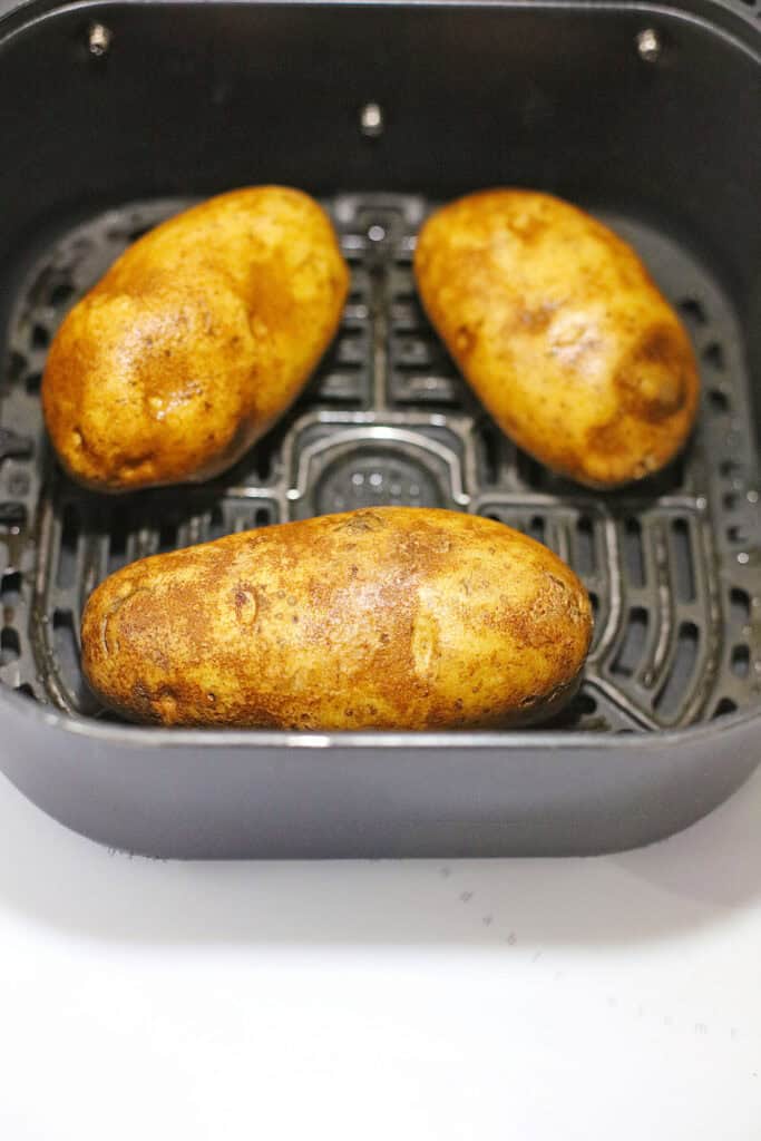 Three whole potatoes are arranged in an air fryer basket, ready to be transformed into delicious chili cheese baked potatoes.