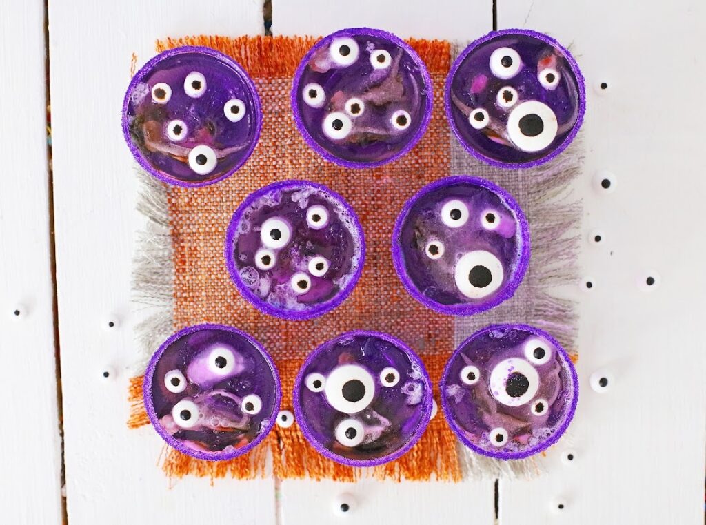 Seven purple cupcakes with white candy eyes sit on an orange mat, resembling Creepy Monster Eye Jello Shots, all on a white wooden surface.