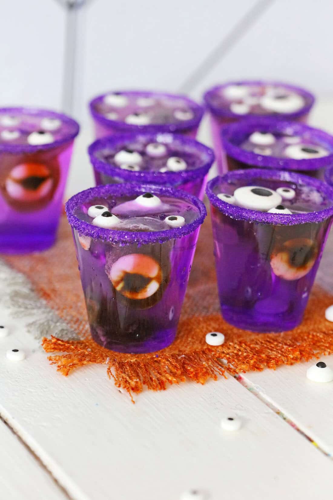 A person pours sugar from a brown paper bag into a measuring cup filled with purple liquid, carefully crafting Monster Eye Jello Shots. A Jell-O packet and other items are visible in the background.