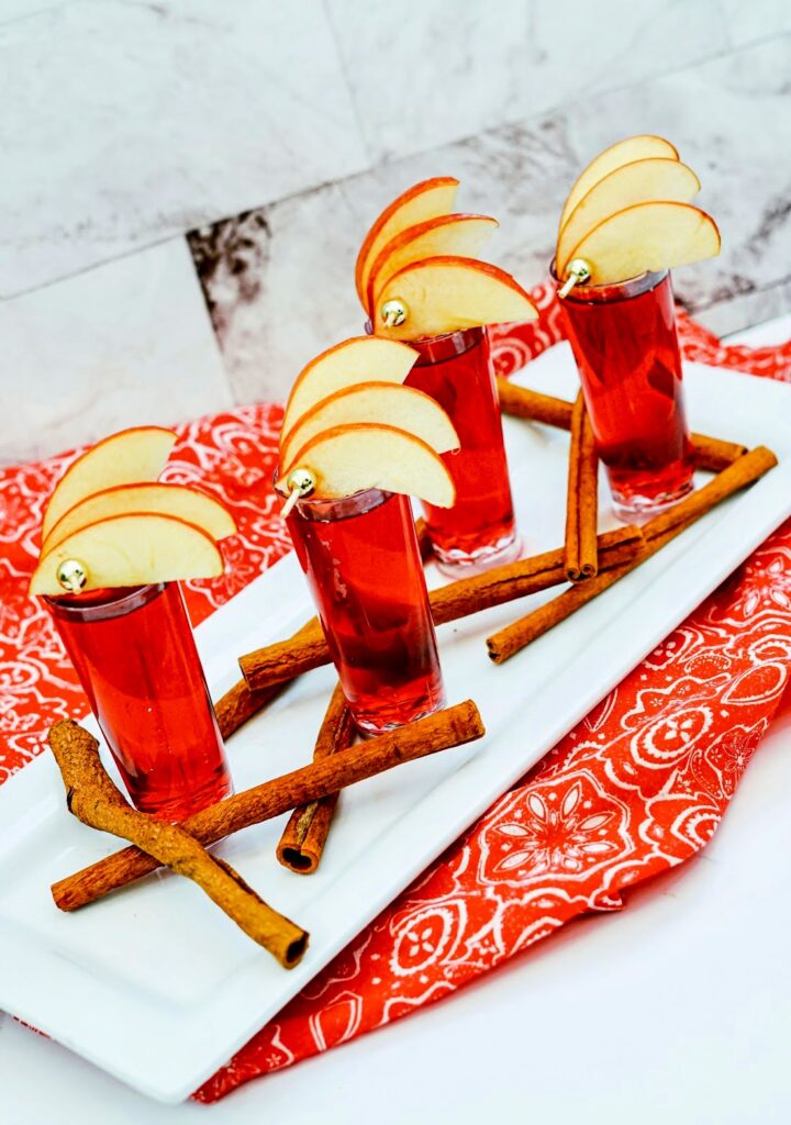 Four red Cran Apple shooters in glasses, garnished with apple slices and cinnamon sticks, arranged on a rectangular white plate with a red patterned cloth underneath.