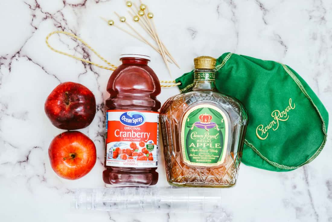 Four red Cran Apple shooters in glasses, garnished with apple slices and cinnamon sticks, arranged on a rectangular white plate with a red patterned cloth underneath.