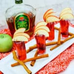 Four Crown Cran Apple Shooters topped with apple slices rest on a tray, surrounded by cinnamon sticks. A bottle of Crown Royal Apple and a green apple complete the scene in the background.