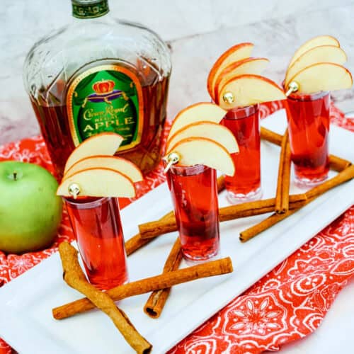 Four Crown Cran Apple Shooters topped with apple slices rest on a tray, surrounded by cinnamon sticks. A bottle of Crown Royal Apple and a green apple complete the scene in the background.