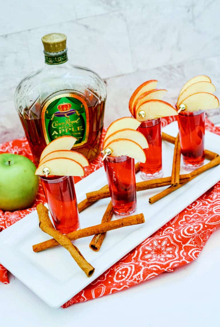 Four Crown Cran Apple Shooters topped with apple slices rest on a tray, surrounded by cinnamon sticks. A bottle of Crown Royal Apple and a green apple complete the scene in the background.