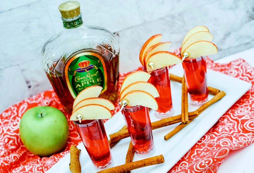 Four apple slice-garnished red cocktails sit on a tray, complemented by cinnamon sticks, a green apple, and a bottle of Cran Apple-flavored whiskey resting on a red patterned cloth.