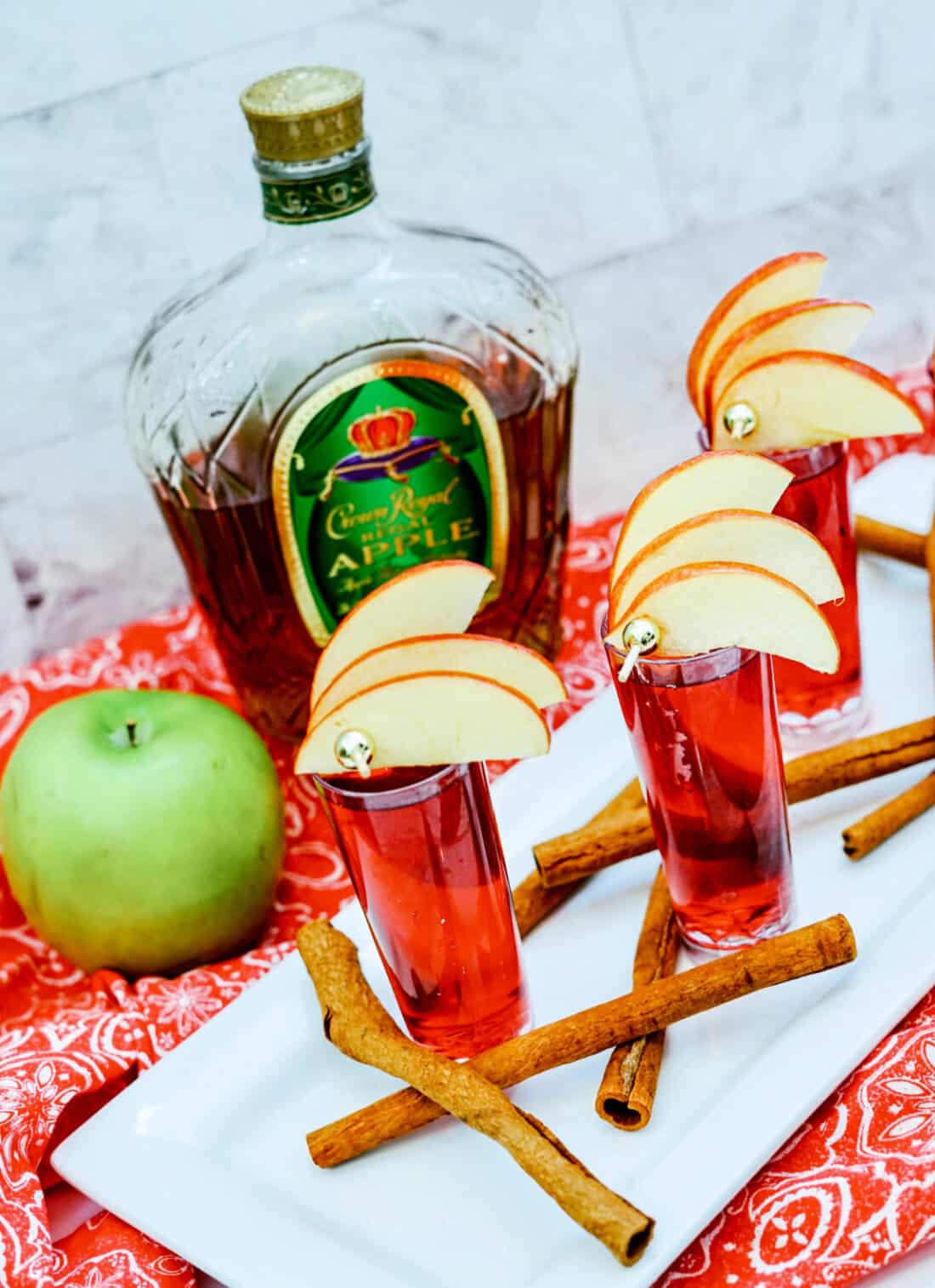 A bottle of Crown Royal Apple whiskey being poured into a glass, accompanied by fresh apples and a green Crown Royal bag on a marble surface, hints at the perfect mix for crafting a refreshing Crown Cran Apple Shooter.