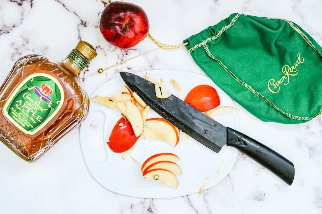 Apple slices and a knife rest on a cutting board beside a Crown Cran Apple Shooter whiskey bottle and a green bag, with a whole apple on a marble surface.