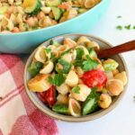 A bowl of lemon tahini pasta salad features shell pasta, chickpeas, cucumbers, tomatoes, and parsley. In the background, a larger dish overflows with more fresh pasta salad. A red checkered cloth adds a rustic touch nearby.