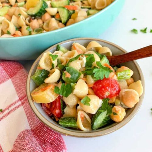 Lemon Tahini Pasta Salad 12 A bowl of lemon tahini pasta salad features shell pasta, chickpeas, cucumbers, tomatoes, and parsley. In the background, a larger dish overflows with more fresh pasta salad. A red checkered cloth adds a rustic touch nearby.