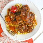 A plate of rice topped with sweet and sour meatballs, garnished with sesame seeds and roasted vegetables, placed on a red and white patterned cloth.