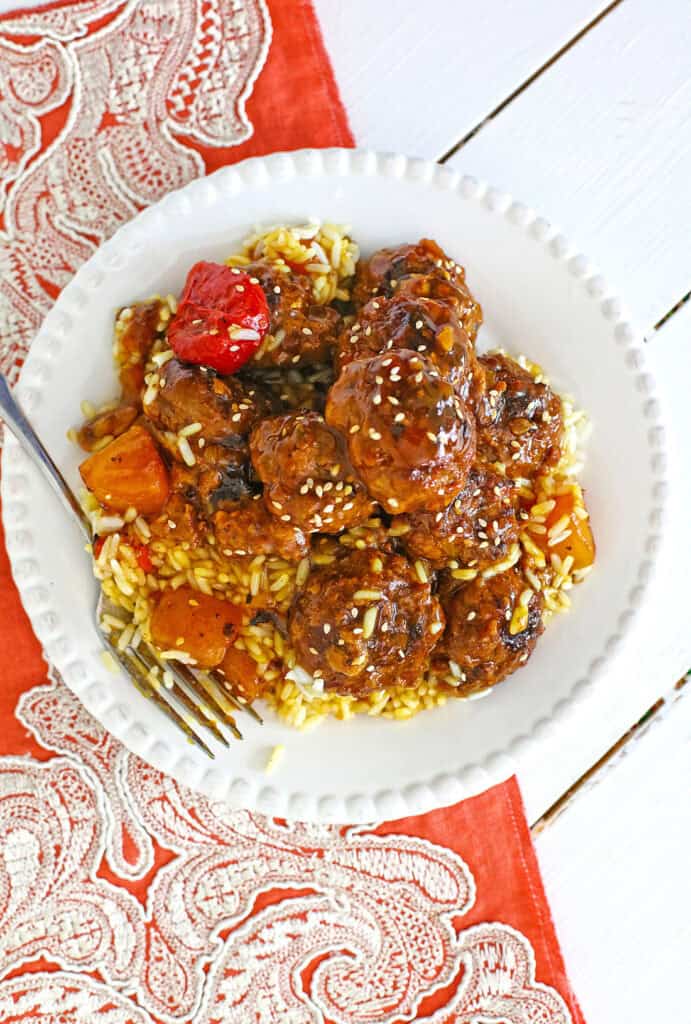 A plate of rice topped with sweet and sour meatballs, garnished with sesame seeds and roasted vegetables, placed on a red and white patterned cloth.