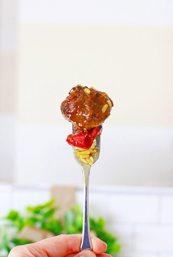Sweet and Sour Meatballs 7 A Sweet and Sour Meatball covered in sauce is held on a fork above a piece of red vegetable and rice. The background is out of focus.