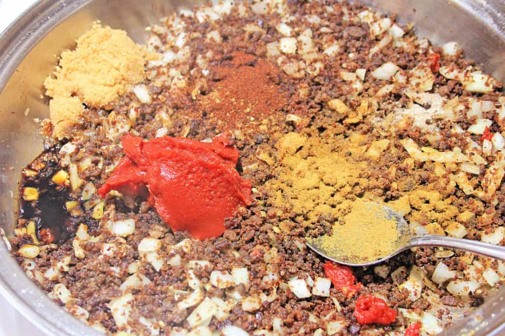 A metal pan contains sautéed ground beef, onions, tomato paste, various powdered spices, and sauce, with a silver spoon resting on the mixture-perfect for topping chili cheese baked potatoes.