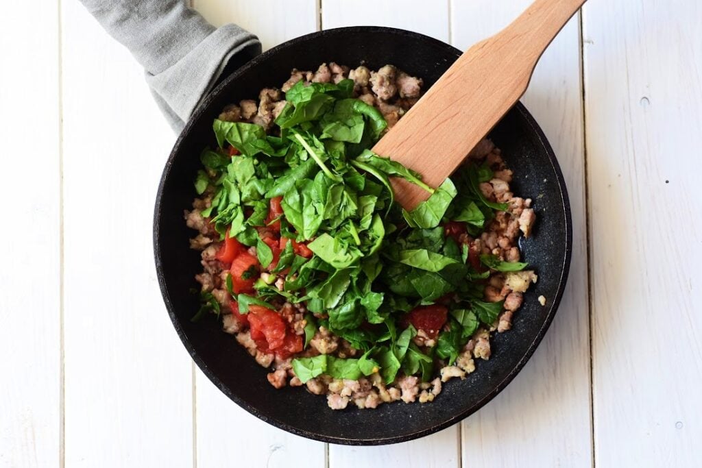 A black skillet on a white surface contains a savory mixture of Italian sausage, cooked ground meat, chopped tomatoes, and fresh spinach. A wooden spatula rests on the skillet's edge, hinting at the perfect filling for ricotta cannelloni. A cloth is nearby.