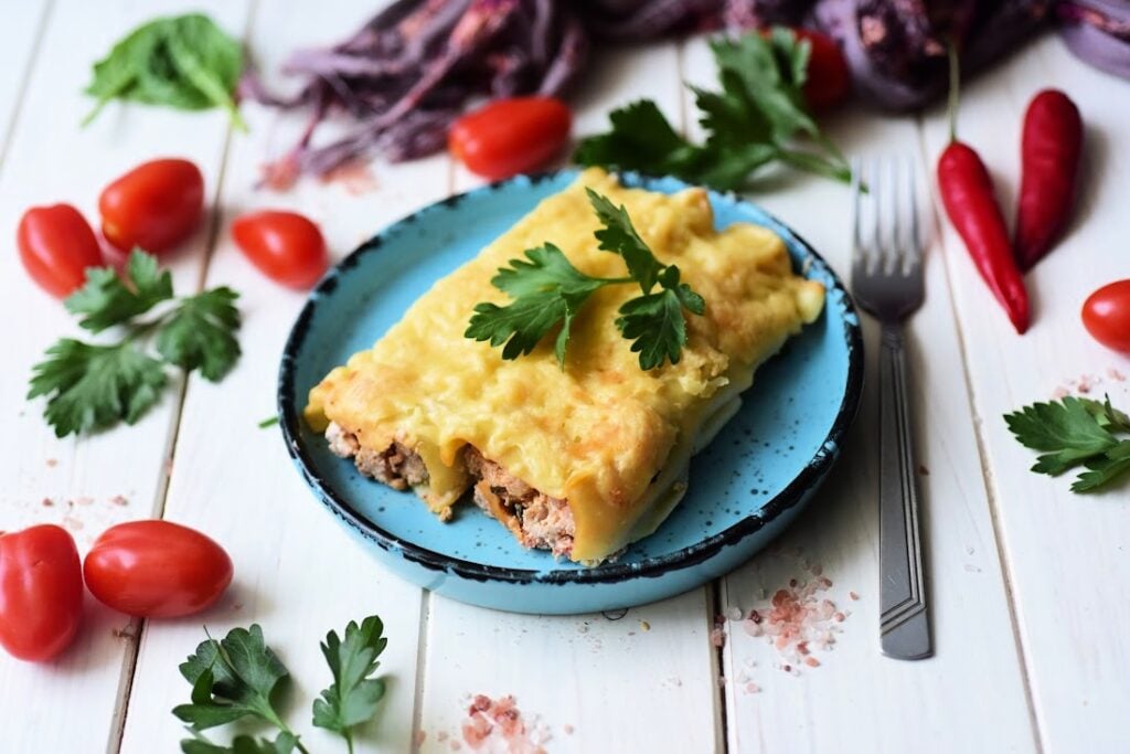 Italian Sausage and Ricotta Cannelloni on a blue plate, garnished with parsley. Surrounded by cherry tomatoes, chili peppers, and parsley leaves on a white wooden surface. Fork nearby.