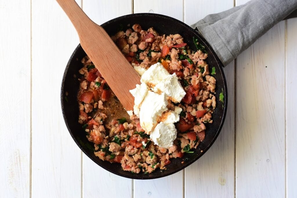 A skillet on a wooden surface brims with an Italian Sausage and Ricotta Cannelloni mixture, featuring savory ground meat and tomatoes topped with ricotta cheese. A wooden spatula rests on the pan's edge, while a gray cloth envelops the handle.
