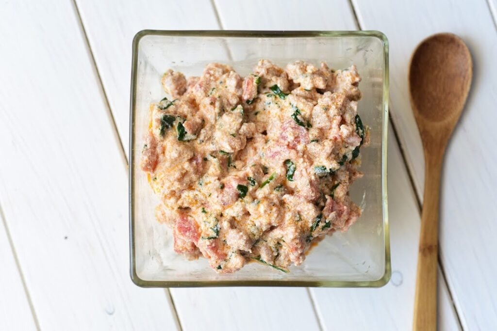 A glass container holds a delightful mixture reminiscent of Italian Sausage and Ricotta Cannelloni, with creamy ingredients, visible herbs, and small red pieces beside a wooden spoon on a white wooden surface.