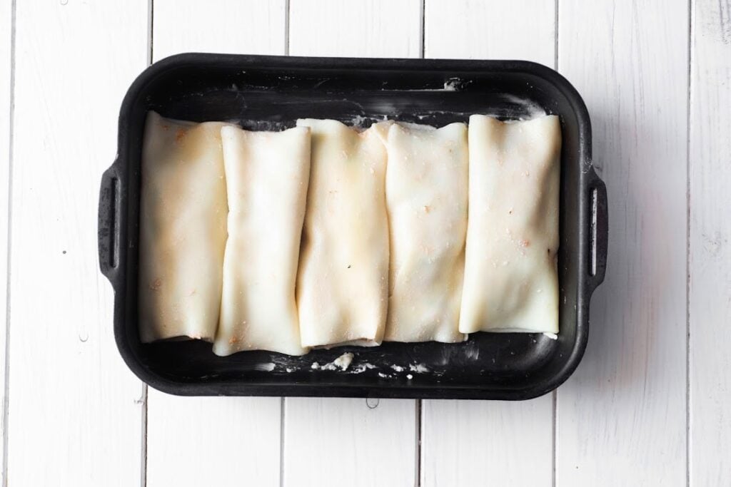 Five uncooked lasagna sheets filled and rolled, resembling Italian Sausage and Ricotta Cannelloni, placed side by side in a black baking tray on a white wooden surface.