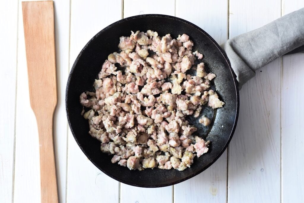 Cooked ground meat sizzles in a black skillet on a white wooden surface, accompanied by a wooden spatula and a gray cloth. This savory base is perfect for crafting Italian Sausage and Ricotta Cannelloni, infusing each roll with rich, hearty flavors.