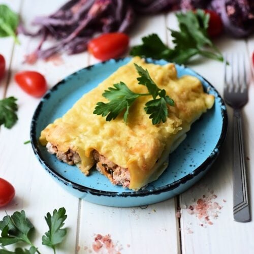 A blue plate showcases Italian Sausage and Ricotta Cannelloni, generously topped with cheese and garnished with parsley. It's artfully surrounded by vibrant cherry tomatoes, fiery chili peppers, and fresh parsley leaves on a white wooden table.