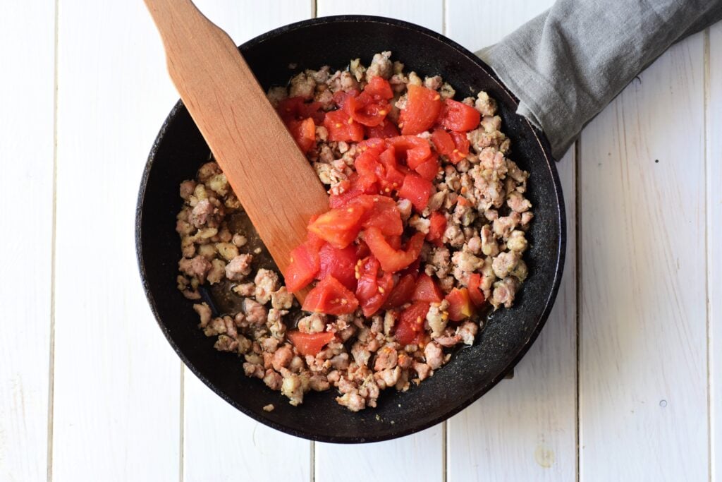 Ground meat and diced tomatoes sizzle in a frying pan, stirred gently with a wooden spatula on a light wooden surface, forming the rich filling for an Italian Sausage and Ricotta Cannelloni delight.