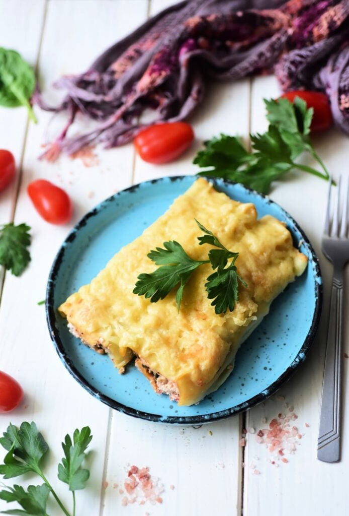 A frying pan containing chopped spinach, diced tomatoes, and cooked ground meat resembles a savory filling for Italian Sausage and Ricotta Cannelloni. A wooden spatula is partially visible, and the pan is placed on a white wooden surface.