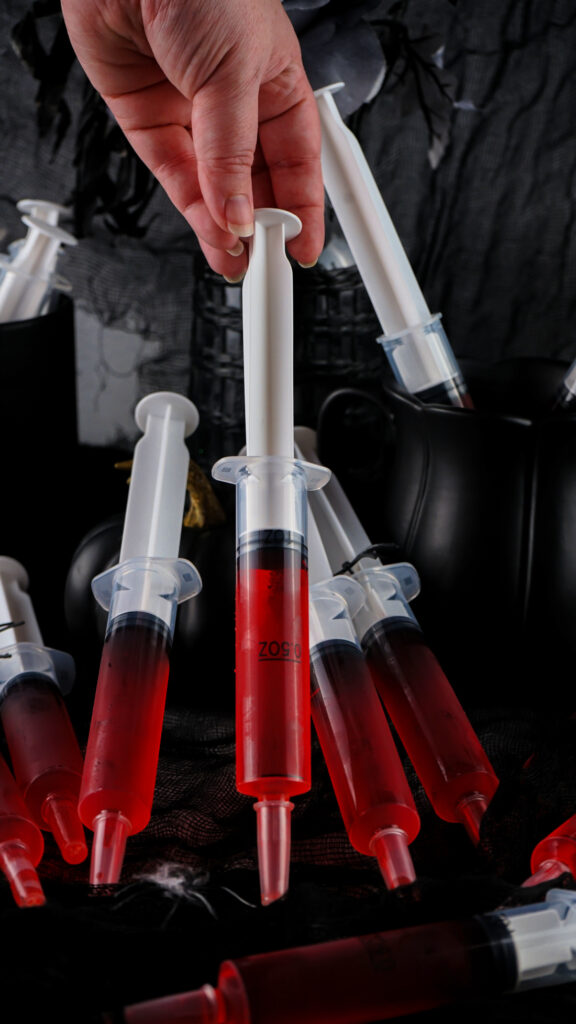 A hand holding a syringe filled with red liquid, reminiscent of playful syringe jello shots, is surrounded by similar syringes on a dark surface, evoking a whimsical yet intriguing atmosphere.