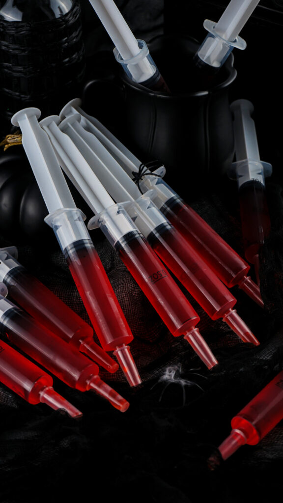 A collection of syringe jello shots filled with red liquid is arranged on a black surface, surrounded by dark fabric and containers.