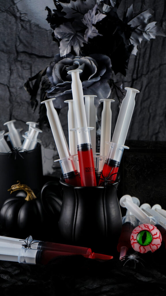 A collection of syringe jello shots filled with red liquid is arranged in a black mug, surrounded by black roses and Halloween-themed decorations.