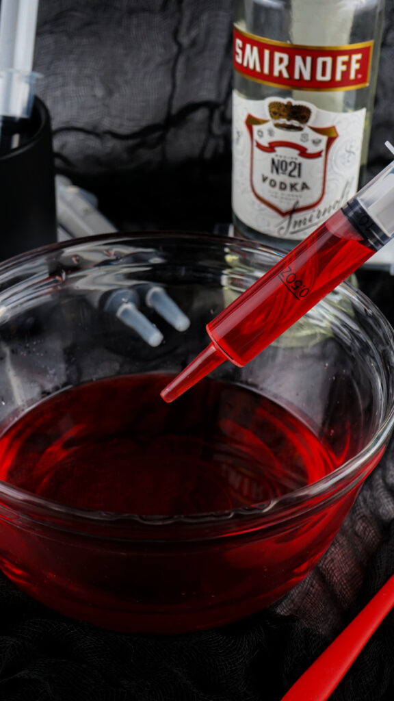 A bowl of red liquid resembling jello shots sits intriguingly with a syringe nearby, accompanied by a bottle of Smirnoff vodka, all set against a dark fabric background.