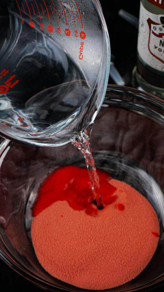 A measuring glass pours clear liquid into a bowl containing red powder, setting the stage for colorful Jello Shots.