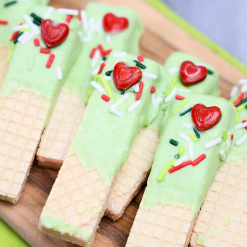Grinch Dipped Wafers: Wafer cookies dipped in green icing, adorned with red heart-shaped candies and colorful sprinkles, artfully arranged on a wooden board.