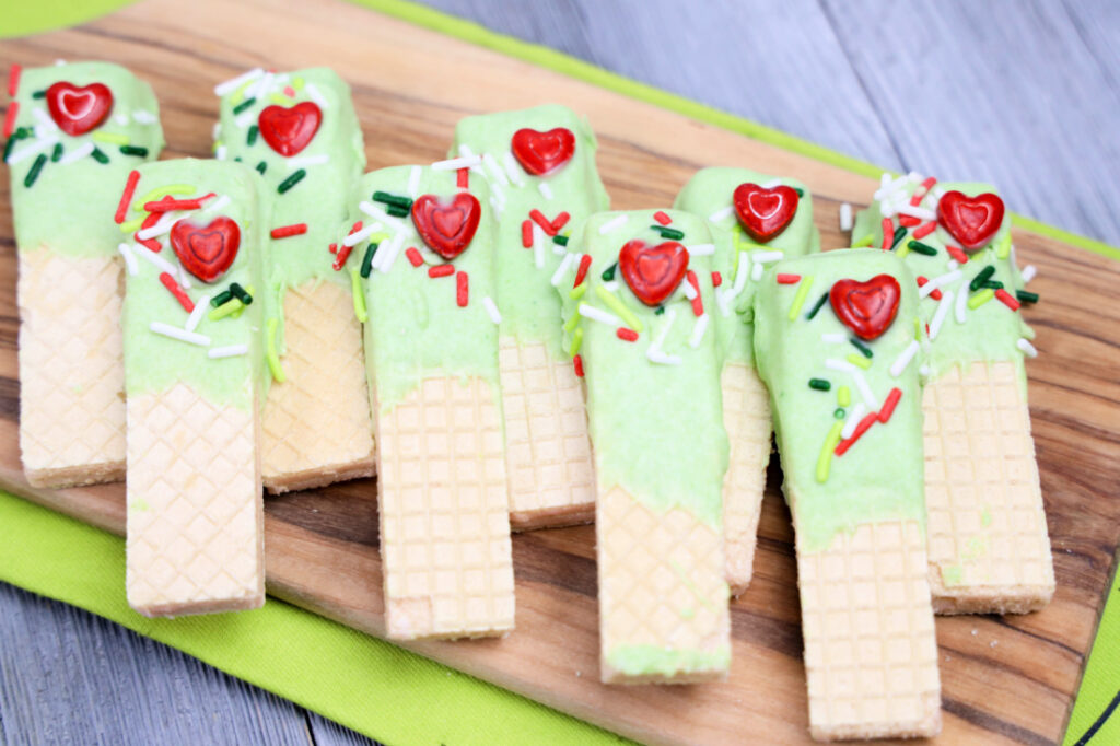 Grinch Dipped Wafer cookies adorned with green icing, heart-shaped candies, and colorful sprinkles are artfully arranged on a wooden tray with a green napkin.