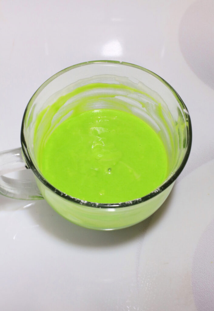 A glass cup filled with bright green liquid sits on a white surface, reminiscent of the vibrant hue found in a Grinch Mug Cake.