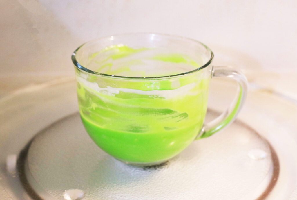 A glass cup with a bright green liquid, reminiscent of a Grinch Mug Cake, sits on a microwave glass plate. The mixture appears partially blended, with some residue clinging to the sides, hinting at the simplicity of an easy mug cake recipe in progress.