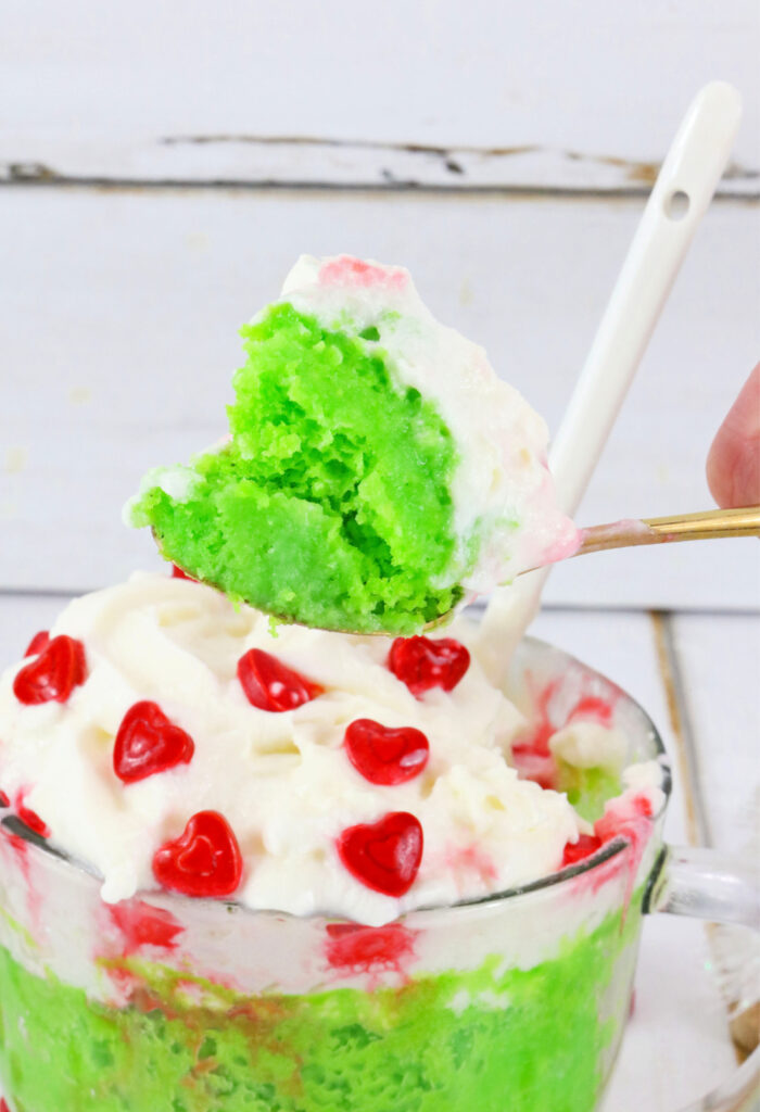 A spoon scoops a Grinch Mug Cake covered in whipped cream with red heart sprinkles from a mug.