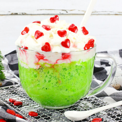A glass mug brimming with a green slushy delight, topped with whipped cream and tiny red heart candies, sits invitingly on a checkered cloth. This Grinch Mug Cake treat is paired perfectly with a spoon for indulgence.
