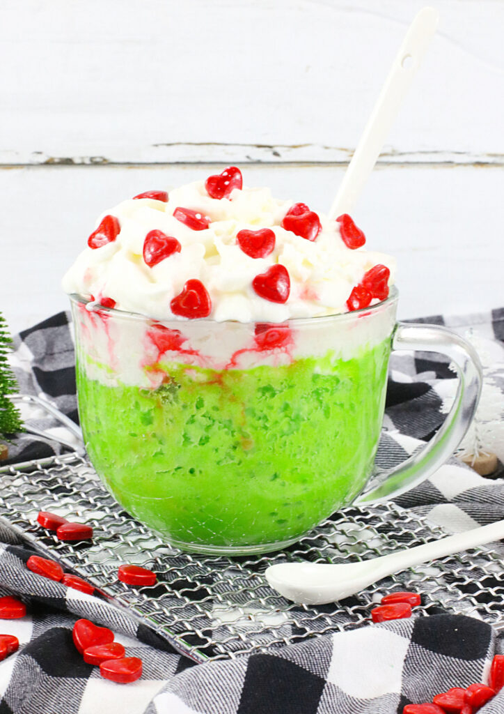 A glass mug brimming with a green slushy delight, topped with whipped cream and tiny red heart candies, sits invitingly on a checkered cloth. This Grinch Mug Cake treat is paired perfectly with a spoon for indulgence.