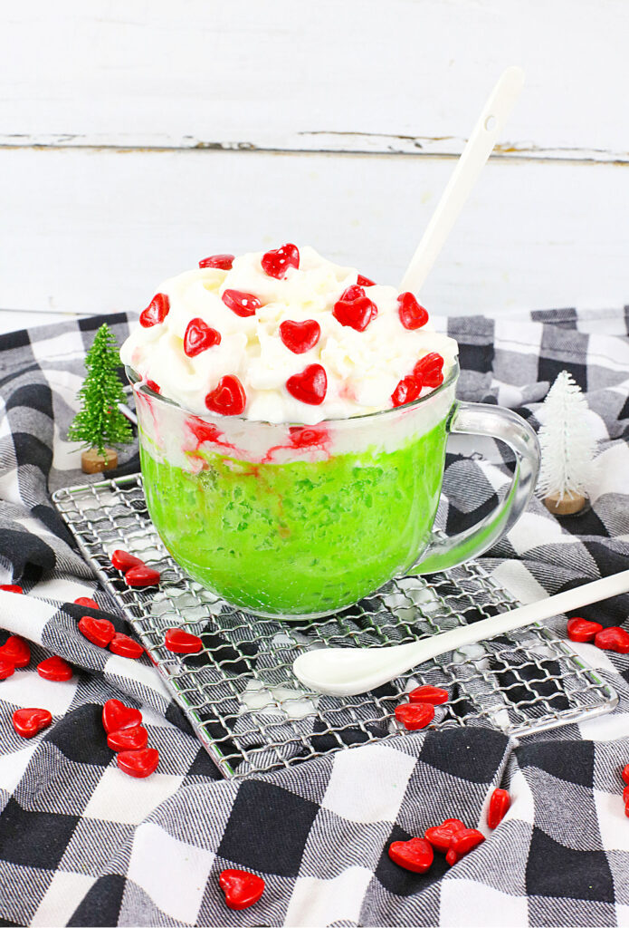 A Grinch Mug Cake sits enticingly on a checkered cloth, its glass mug filled with green dessert and crowned with white whipped cream. Red heart-shaped candies add a whimsical touch, while small decorative trees stand nearby, completing the festive scene.