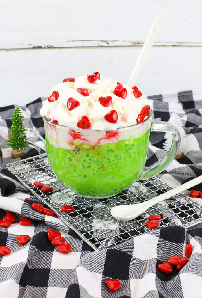 Clear mug with green dessert like a Grinch Mug Cake, topped with whipped cream and heart-shaped red candies on a black and white checkered cloth. A white spoon rests beside the mug, while a small tree decoration adds charm to this easy mug cake recipe scene.