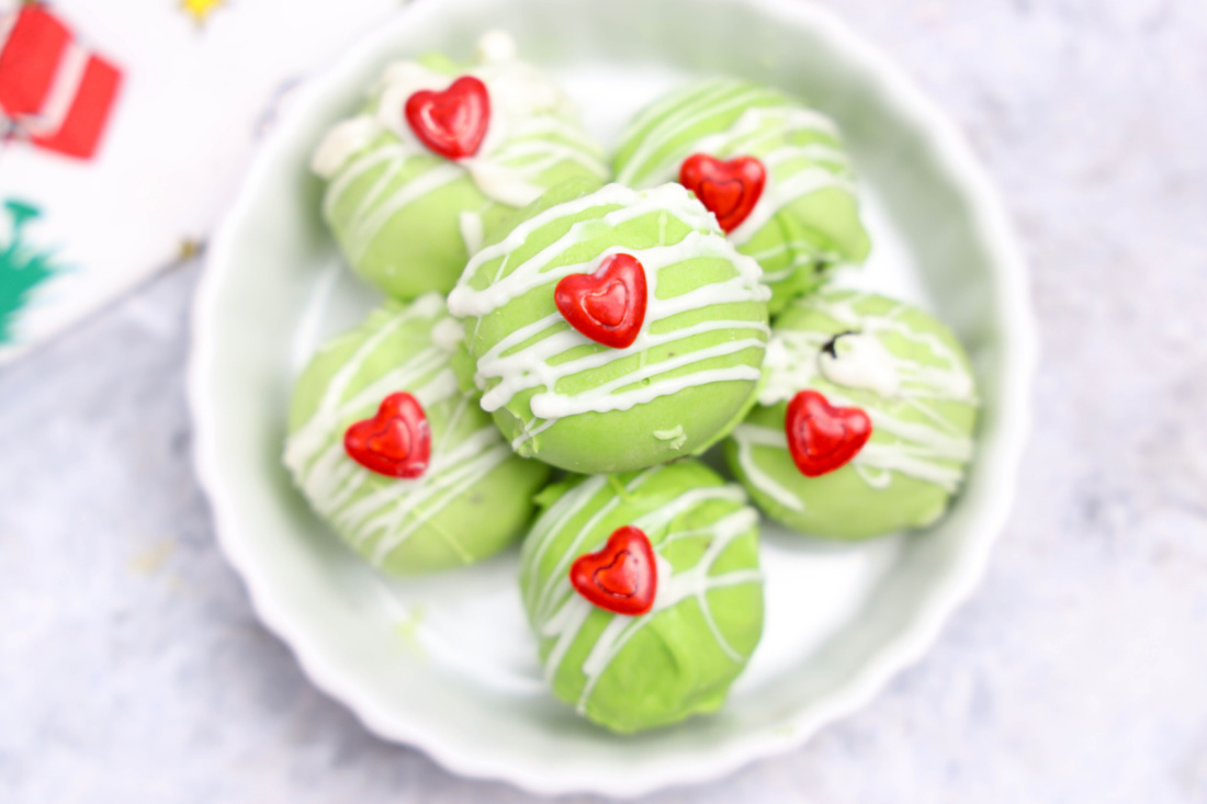 A plate of Grinch Oreo Balls drizzled with white icing, each topped with a red heart decoration, makes for a festive addition to your Christmas recipes collection. 